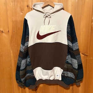 May include: A Nike hooded sweatshirt featuring a cream-colored upper chest and hood, a brown mid-section, and a cream-colored kangaroo pocket. The sleeves are made of a multi-colored knit fabric with horizontal stripes in blue, brown, and grey.