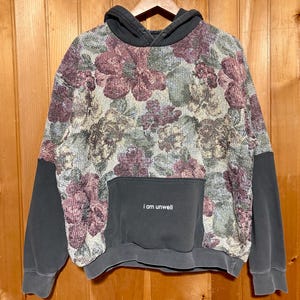 Könnte beinhalten: Ein anthrazitfarbener Kapuzenpullover mit einem floralen Wandteppichmuster in Burgunderrot, Salbeigrün und Creme auf der Vorder- und Rückseite. Das Kleidungsstück verfügt über eine anthrazitfarbene Kängurutasche mit dem Aufdruck i am unwell.