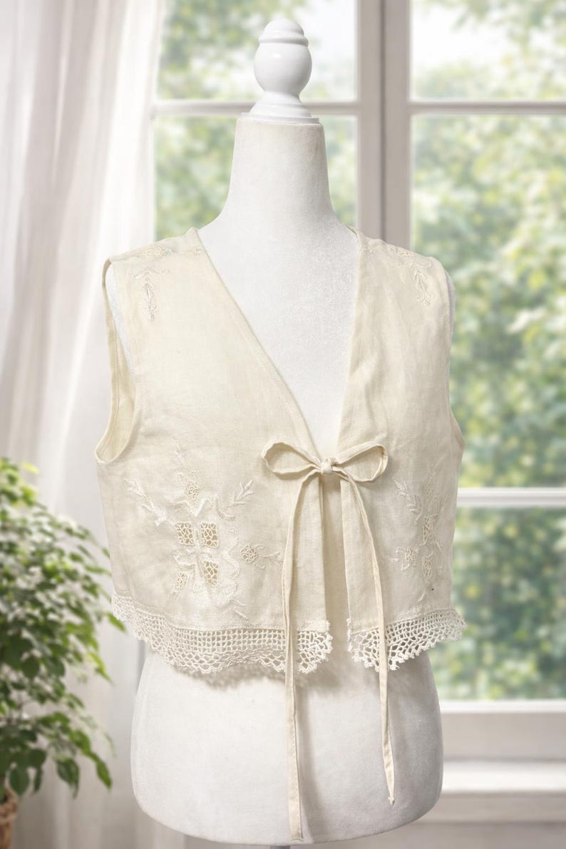 Pu&ograve; includere: Gilet senza maniche color avorio con scollo a V e chiusura con fiocco. Il gilet presenta delicati ricami floreali e un bordo in pizzo all'uncinetto sul bordo inferiore. L'indumento &egrave; esposto su un manichino.