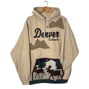 May include: A tan-coloured hoodie featuring the text "Denver Colorado" in black script, with a mountain graphic. The front pocket has a forest scene with two moose. The hoodie has a drawstring hood and a green hem.