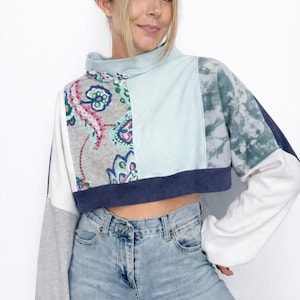 Puede incluir: Un top corto de patchwork con cuello alto y mangas largas. El top presenta una mezcla de estampados y colores, incluyendo cachemir, teñido anudado y paneles lisos en azul, blanco y gris. Las mangas son una mezcla de blanco y gris.