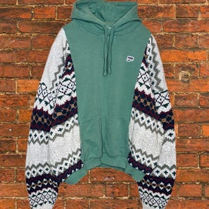Puede incluir: Una sudadera con capucha verde con mangas estampadas. El cuerpo de la sudadera es verde liso, mientras que las mangas presentan un diseño geométrico blanco, gris, burdeos y azul marino. La sudadera tiene un bolsillo delantero y una capucha con cordón.