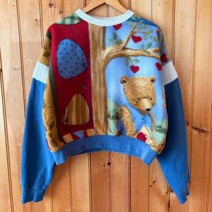 Puede incluir: Una sudadera vintage de cuello redondo con un diseño caprichoso de oso y árbol. La sudadera tiene mangas azules y un cuello y dobladillo azul claro. El panel frontal tiene un diseño de patchwork con un oso de peluche leyendo un libro debajo de un árbol con hojas en forma de corazón.