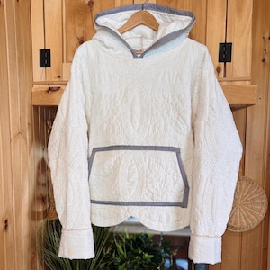May include: Cream-colored quilted hoodie with a front pocket and hood. The hoodie features a blue and white striped trim on the hood, pocket, and cuffs. The fabric has a detailed, textured pattern. The hoodie is hanging on a wooden hanger.