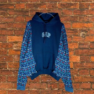 May include: A navy blue hoodie with the GAP logo in white. The sleeves feature a patterned design in blue, red, and white. The hoodie is displayed against a brick wall background.