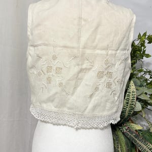 Pu&ograve; includere: Gilet color crema con ricami floreali e un delicato bordo in pizzo. Il gilet &egrave; esposto su un manichino, mettendo in risalto i suoi dettagli intricati e l'estetica vintage. Un capo senza tempo.
