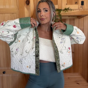 Könnte beinhalten: Cremefarbene Steppjacke mit floraler Stickerei und grünen Zierleisten. Die Jacke hat eine Knopfleiste und wird mit tealfarbenen Sporthosen kombiniert. Das Design der Jacke zeigt ein zartes Muster aus Blumen und Blättern.