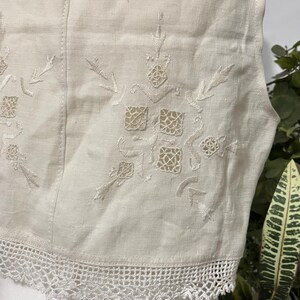 Pu&ograve; includere: Primo piano di un gilet color crema con intricati motivi floreali e geometrici ricamati. Il gilet presenta un delicato bordo in pizzo bianco sul bordo inferiore. Il tessuto sembra essere lino o una fibra naturale simile.