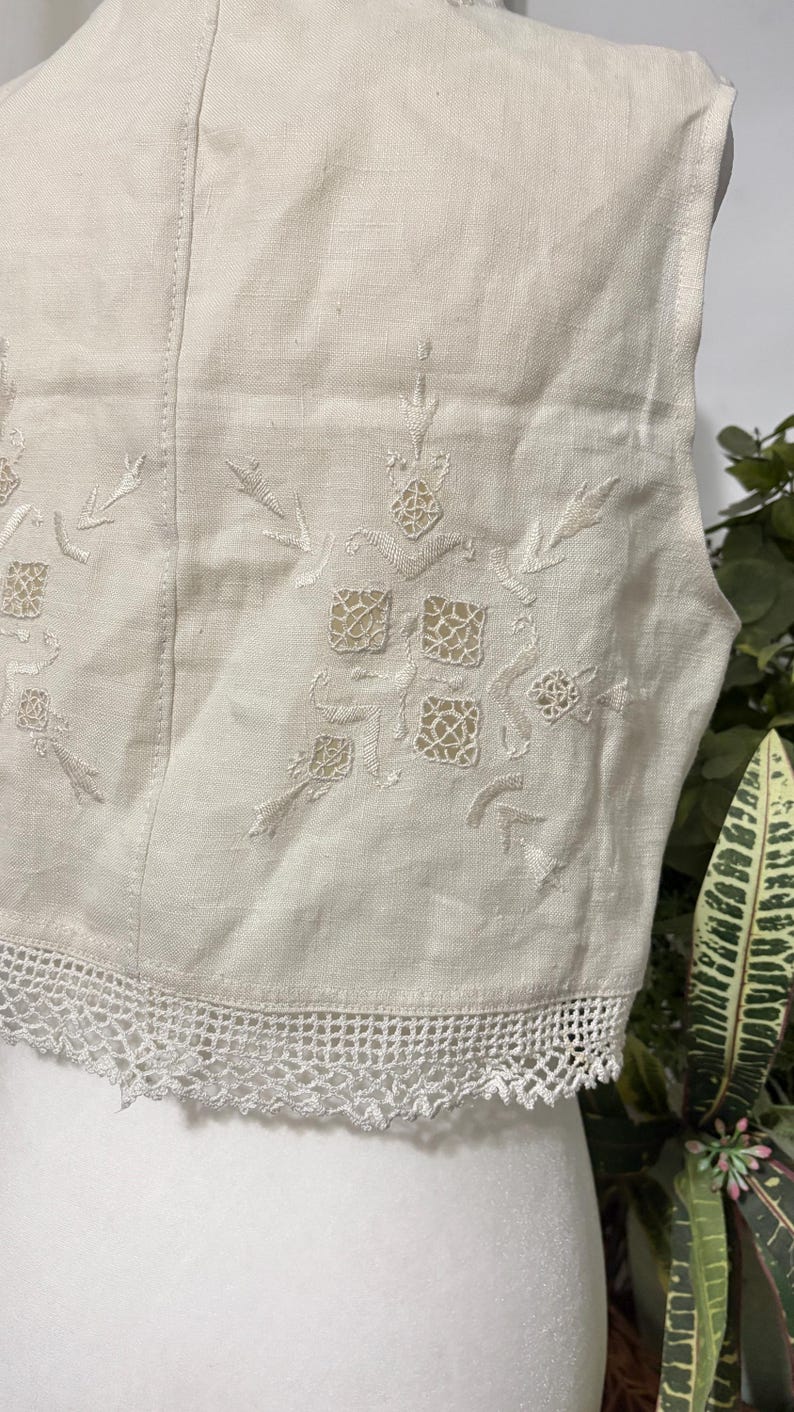 Pu&ograve; includere: Primo piano di un gilet color crema con intricati motivi floreali e geometrici ricamati. Il gilet presenta un delicato bordo in pizzo bianco sul bordo inferiore. Il tessuto sembra essere lino o una fibra naturale simile.