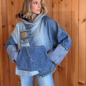 May include: A patchwork denim hoodie in shades of blue and grey. The hoodie features a kangaroo pocket, a front pocket with a brown leather patch, and a grey hood. The sleeves have wide cuffs. The garment is worn over blue jeans.