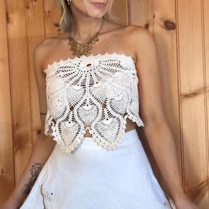 Könnte beinhalten: Cremefarbenes, handgefertigtes Häkeltop mit Muschelkante. Das Top hat ein detailliertes, offenes Design. Das Model trägt eine goldene Halskette und einen weißen Rock. Der Hintergrund ist eine Holzwand.