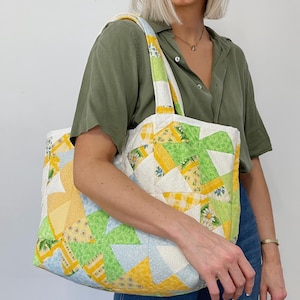 Könnte beinhalten: Eine große Patchwork-Tragetasche mit gelben, grünen und blauen Stoffquadraten. Die Tasche hat einen weißen Hintergrund und ein Blumenmuster. Die Tasche wird von einer Person getragen, die ein grünes Hemd und eine blaue Jeans trägt.