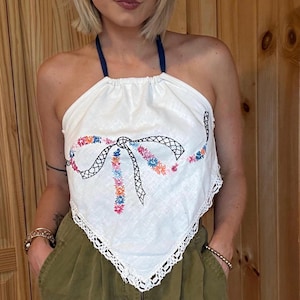 May include: White halter top with a floral embroidered bow design and a navy blue halter strap. The top has a lace trim along the bottom edge. The floral embroidery includes pink, orange, blue, and yellow colors.