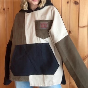 Może przedstawiać: Bluza z kapturem typu patchwork w odcieniach czerni, brązu i beżu. Bluza posiada dużą przednią kieszeń i małą kieszeń na piersi. Rękawy są w jednolitym brązowym kolorze, a kaptur jest czarny.