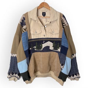 May include: A patchwork sweater with a tan denim collar and two front pockets. The sweater features a landscape design with trees and a crescent moon. The sleeves have a patterned knit design. The sweater is hanging on a wooden hanger.