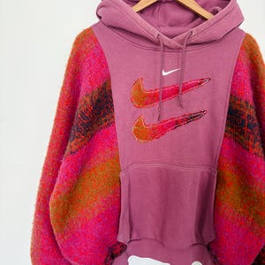 Puede incluir: Una sudadera con capucha malva con bolsillo de canguro y capucha, con dos logotipos de Nike bordados. Las mangas son de una tela vibrante y texturizada con rayas rosas, naranjas y negras. La sudadera está colgada en una percha de madera.