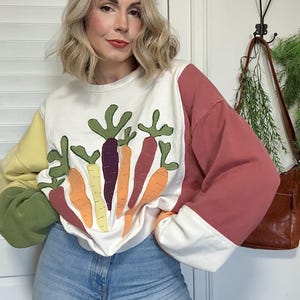 Puede incluir: Una sudadera blanca con un diseño de zanahorias coloridas. Las mangas son de bloques de color amarillo, verde y rojo. La persona lleva vaqueros azules.