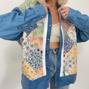 Op de afbeelding: Een blauwe denimjas met een patchwork-ontwerp met bloemen- en geometrische patronen. De jas heeft een knoopsluiting aan de voorkant en lange mouwen.