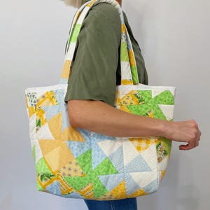 May include: A large patchwork tote bag with a white background and colorful geometric patterns in shades of yellow, green, and blue. The bag has two long straps and is made of quilted fabric.