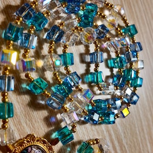 May include: A close-up of a rosary featuring blue, clear, and gold beads. The beads are square-cut and connected with gold-coloured wire. A gold-coloured pendant with a religious image is at the bottom. The rosary is set against a wooden background.