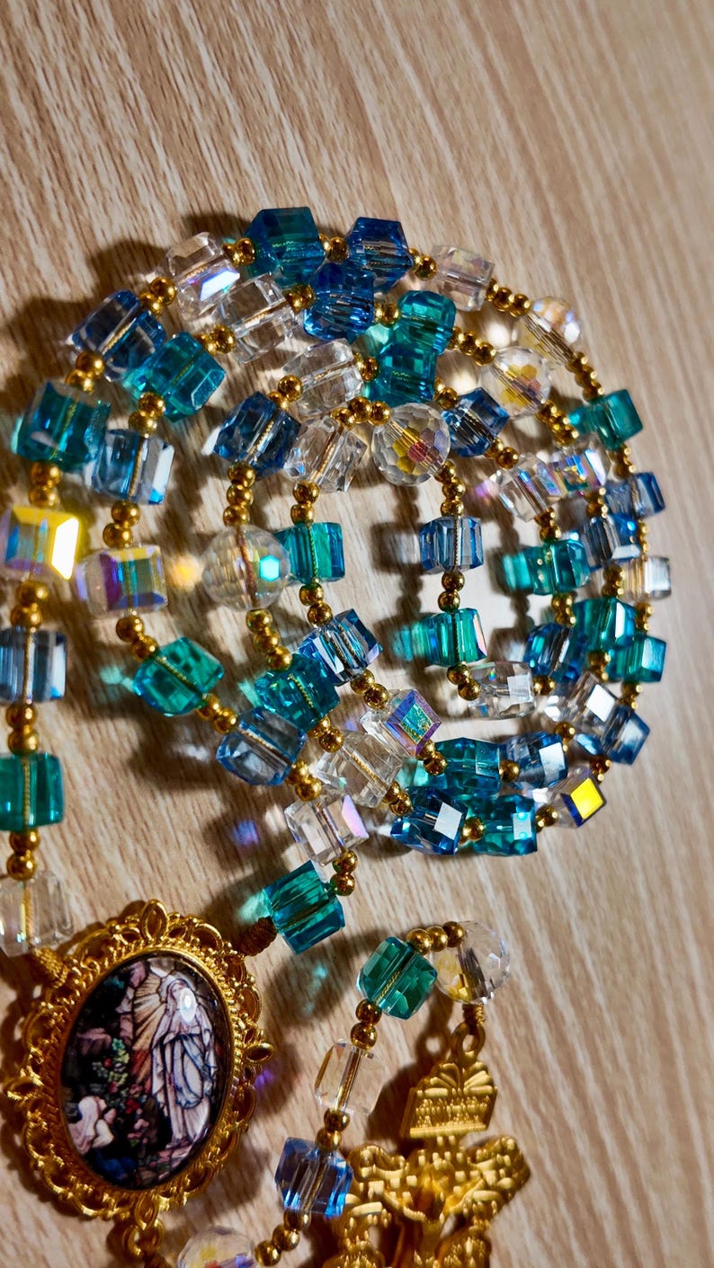 May include: A close-up of a rosary featuring blue, clear, and gold beads. The beads are square-cut and connected with gold-coloured wire. A gold-coloured pendant with a religious image is at the bottom. The rosary is set against a wooden background.