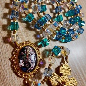 May include: A rosary featuring blue, clear, and gold beads. The rosary includes a gold-coloured crucifix and a medallion with a religious image. The beads are a mix of faceted and round shapes.