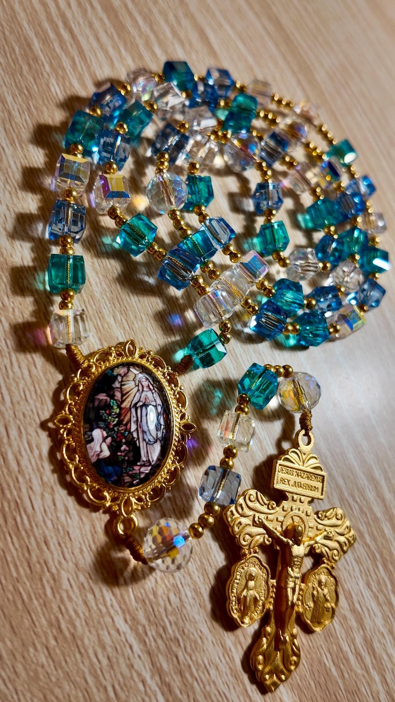 May include: A rosary featuring blue, clear, and gold beads. The rosary includes a gold-coloured crucifix and a medallion with a religious image. The beads are a mix of faceted and round shapes.