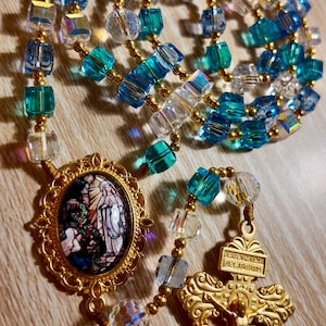 May include: A rosary featuring blue, clear, and gold beads. It includes a gold crucifix and a gold pendant with a religious image. The beads are a mix of faceted and round shapes.