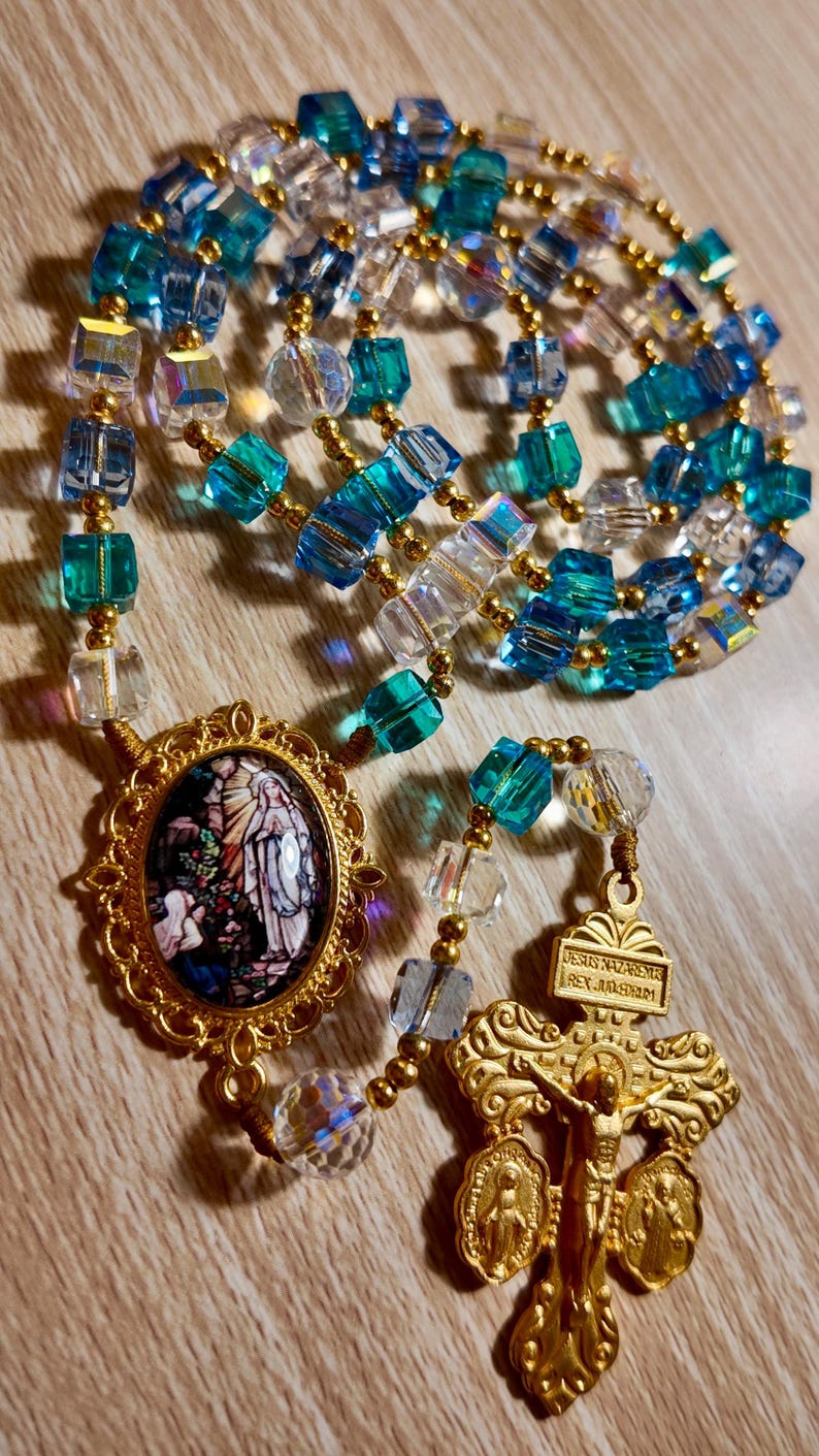 May include: A rosary featuring blue, clear, and gold beads. It includes a gold crucifix and a gold pendant with a religious image. The beads are a mix of faceted and round shapes.