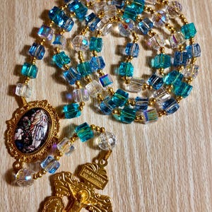 May include: A rosary featuring blue, clear, and gold beads. The beads are square and faceted, with gold-coloured spacers. The rosary has a gold-coloured crucifix and a gold-coloured oval pendant with a religious image.