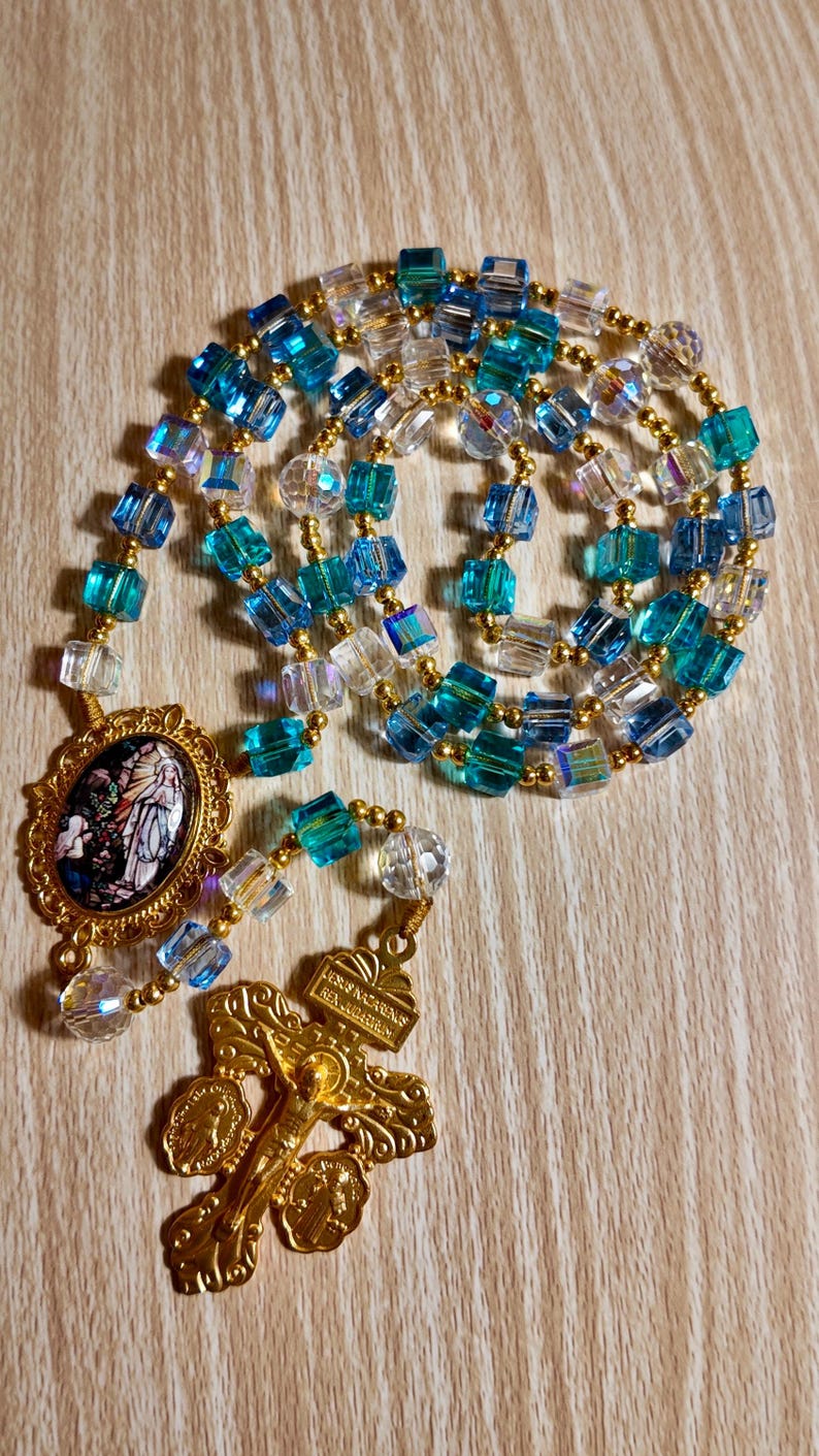 May include: A rosary featuring blue, clear, and gold beads. The beads are square and faceted, with gold-coloured spacers. The rosary has a gold-coloured crucifix and a gold-coloured oval pendant with a religious image.