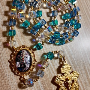 May include: A rosary featuring blue, clear, and gold beads. The beads are a mix of square and round shapes. The rosary includes a gold-coloured crucifix and a pendant with a religious image.