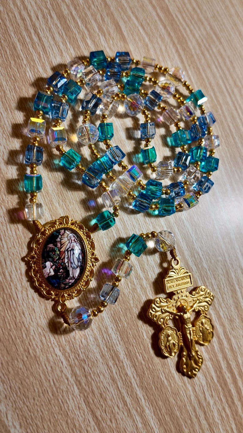 May include: A rosary featuring blue, clear, and gold beads. The beads are a mix of square and round shapes. The rosary includes a gold-coloured crucifix and a pendant with a religious image.