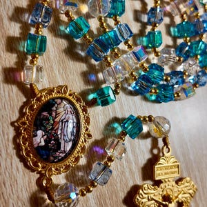 May include: A rosary featuring blue, clear, and gold beads. The beads are a mix of faceted cubes and round shapes. A gold pendant displays a religious image, and a gold crucifix hangs at the bottom.