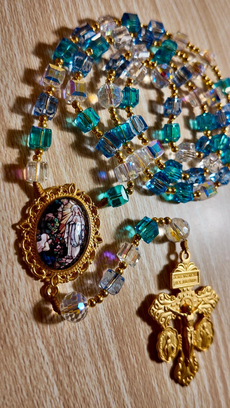 May include: A rosary featuring blue, clear, and gold beads. The beads are a mix of faceted cubes and round shapes. A gold pendant displays a religious image, and a gold crucifix hangs at the bottom.