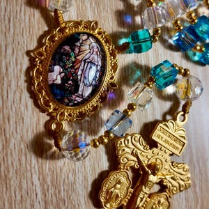 May include: A gold-toned rosary with blue and clear crystal beads. The rosary features a detailed crucifix and a pendant with a religious image. The beads are connected by gold-coloured wire.