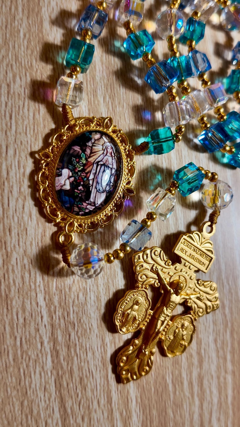 May include: A gold-toned rosary with blue and clear crystal beads. The rosary features a detailed crucifix and a pendant with a religious image. The beads are connected by gold-coloured wire.
