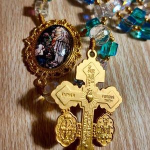 May include: A gold rosary with blue and clear beads. The rosary features a cross pendant with the words "Father" and "Forgive Them." An oval pendant with a stained-glass image is also present.