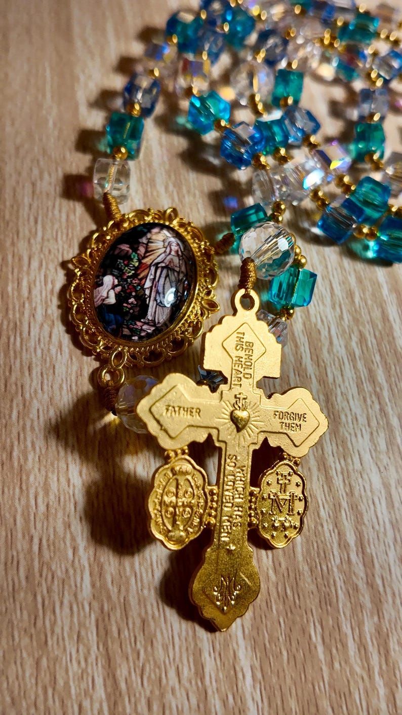 May include: A gold rosary with blue and clear beads. The rosary features a cross pendant with the words "Father" and "Forgive Them." An oval pendant with a stained-glass image is also present.