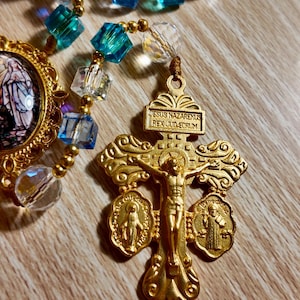 May include: A gold rosary with blue and clear beads. The rosary features a crucifix with the inscription "JESUS NAZARENUS REX JUD-EORUM." Additional religious medallions are attached to the crucifix.