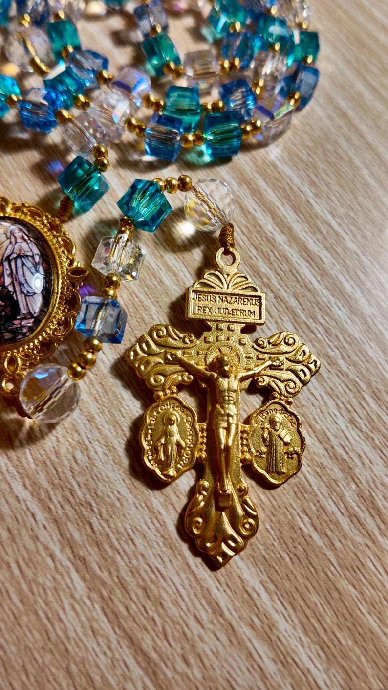 May include: A gold rosary with blue and clear beads. The rosary features a crucifix with the inscription "JESUS NAZARENUS REX JUD-EORUM." Additional religious medallions are attached to the crucifix.