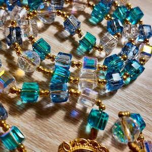 May include: A necklace with a gold pendant and a stained-glass design, linked to strands of clear, blue, and teal cube-shaped beads. The beads are interspersed with gold-coloured beads. The necklace is displayed on a wooden surface.