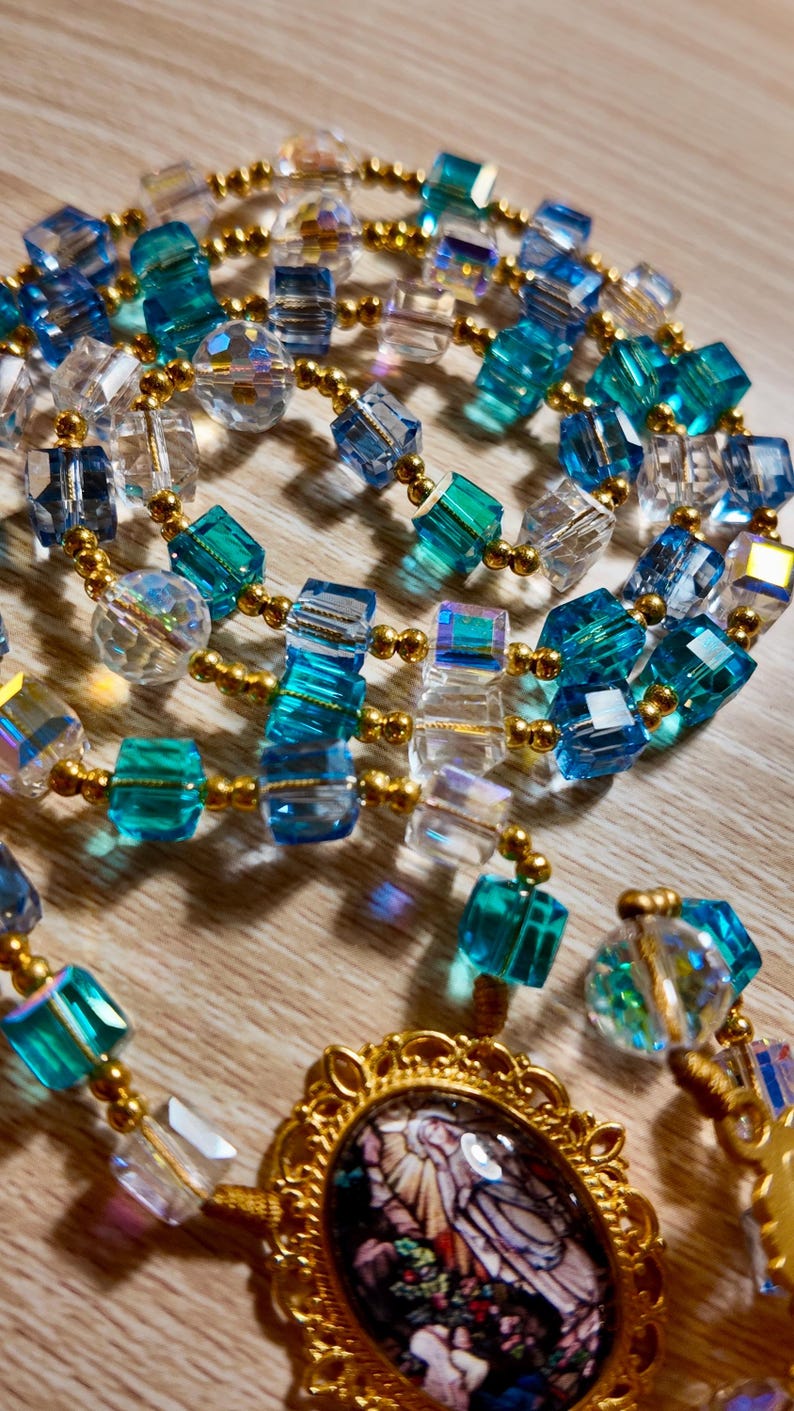 May include: A necklace with a gold pendant and a stained-glass design, linked to strands of clear, blue, and teal cube-shaped beads. The beads are interspersed with gold-coloured beads. The necklace is displayed on a wooden surface.