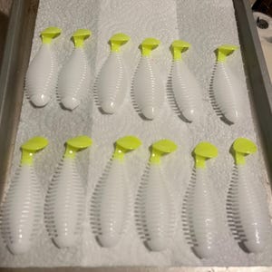 May include: Twelve white plastic fishing lures arranged in two rows of six. Each lure features a ribbed, elongated body and a bright neon yellow paddle tail. The lures are positioned on a white paper towel inside a metal baking sheet.