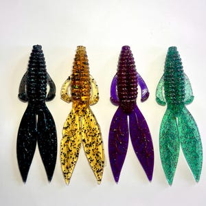May include: Four soft plastic fishing lures arranged in a row. Each lure features a ribbed body, two side appendages, and a split tail. The colors from left to right are black with blue glitter, amber with black glitter, purple with red glitter, and green with red glitter.