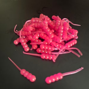 May include: A pile of pink plastic fishing bait, also known as grubs, with a few individual grubs laying on a black surface.