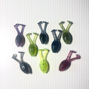 May include: Assortment of eight frog-shaped fishing lures in various colors, including green, purple, black, and pink. The lures have speckled patterns and are designed to resemble frogs.