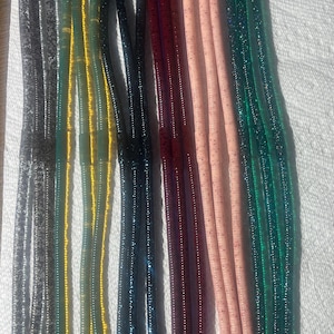 May include: An assortment of soft plastic fishing lures in various colors, including gray, yellow, dark blue, burgundy, and pink, with glitter accents. The lures are arranged in parallel rows on a white surface, showcasing their different shades and textures.