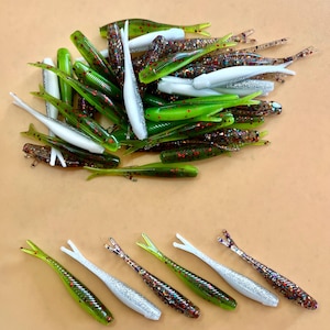 May include: A collection of soft plastic fishing lures shaped like small fish. The lures come in various colors including solid white, translucent green with red glitter, and dark brown with multi-colored glitter, all featuring split tails.