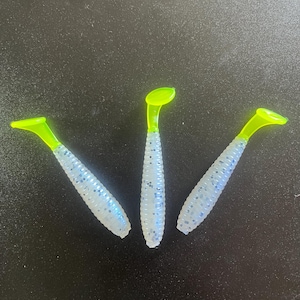 May include: Three soft plastic fishing lures. Each lure has a clear body with blue flecks and a bright green paddle tail. The lures are arranged in a fan shape against a dark background.